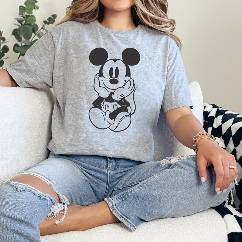 Peut inclure: Un t-shirt gris avec un dessin noir et blanc de Mickey Mouse. Le personnage de dessin anim&eacute; est assis, les mains sous le menton. Le t-shirt est port&eacute; avec un jean bleu.