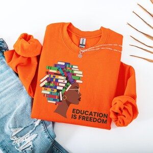 Puede incluir: Sudadera naranja brillante con un gr&aacute;fico del perfil de una mujer con libros formando su cabello y el texto "EDUCATION IS FREEDOM". Un collar plateado est&aacute; sobre la sudadera. La sudadera est&aacute; doblada junto a unos vaqueros azules.