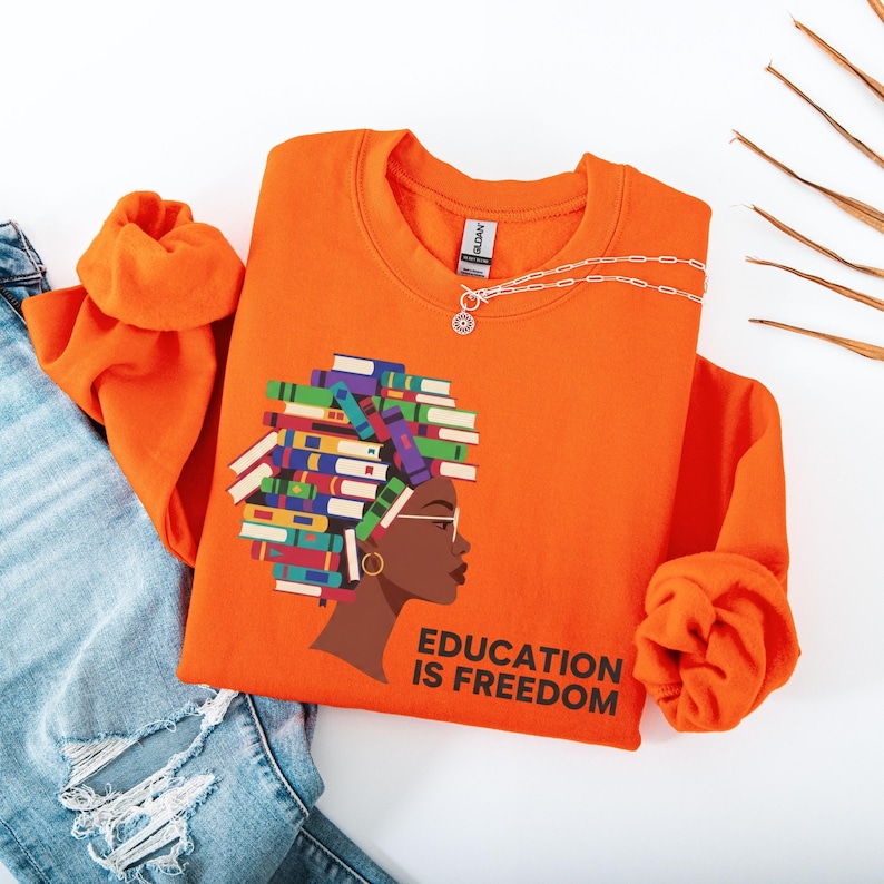 Puede incluir: Sudadera naranja brillante con un gr&aacute;fico del perfil de una mujer con libros formando su cabello y el texto "EDUCATION IS FREEDOM". Un collar plateado est&aacute; sobre la sudadera. La sudadera est&aacute; doblada junto a unos vaqueros azules.