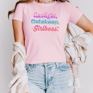 Może przedstawiać: Jasnor&oacute;żowa koszulka z napisem "Gaslight, Gatekeep, Girlboss" w kolorowej, retro czcionce. Koszulka z kr&oacute;tkim rękawem jest wykonana z miękkiego materiału. Modelka ma na sobie niebieskie dżinsy.