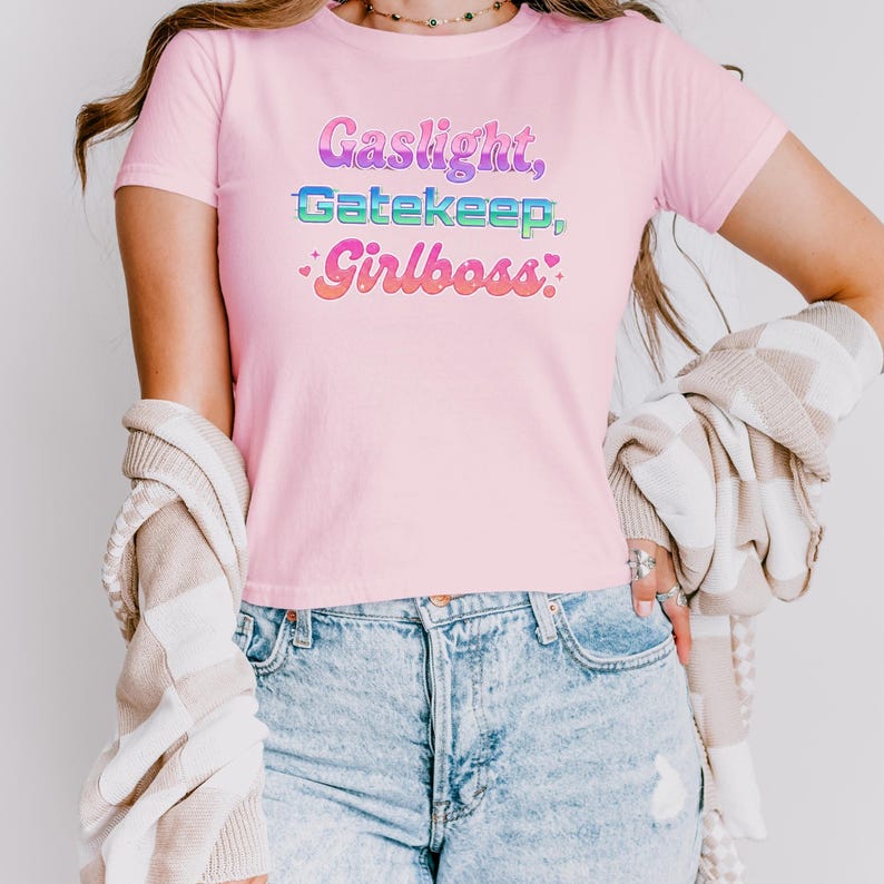 Może przedstawiać: Jasnor&oacute;żowa koszulka z napisem "Gaslight, Gatekeep, Girlboss" w kolorowej, retro czcionce. Koszulka z kr&oacute;tkim rękawem jest wykonana z miękkiego materiału. Modelka ma na sobie niebieskie dżinsy.