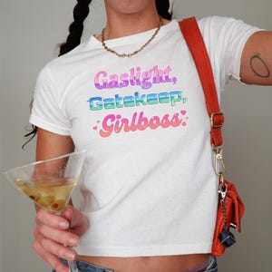 Może przedstawiać: Biała koszulka z napisem "Gaslight, Gatekeep, Girlboss" w kolorowych, gradientowych literach. Koszulka jest połączona ze złotym łańcuszkiem i pomarańczową torbą na ramię. W dłoni trzymany jest kieliszek do martini z oliwkami.