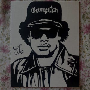 Puede incluir: Pintura de retrato en blanco y negro de una persona con gafas de sol y un sombrero con la palabra "Compton". La pintura tiene una firma en la esquina inferior izquierda.
