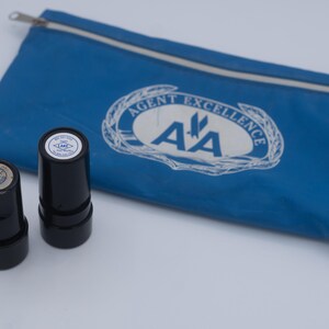 May include: Two black stampers with circular imprints and a blue zippered pouch. The pouch has the text "AGENT EXCELLENCE" and the letters "AA" in a white oval. The stampers have circular imprints.