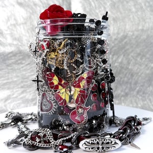 Puede incluir: Un tarro de cristal transparente lleno de collares, pulseras y abalorios en plata, oro, negro y rojo. Una rosa roja está encima del tarro. Un gran colgante de mariposa y un colgante de guitarra son visibles.
