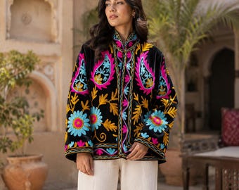 Puede incluir: Una chaqueta de terciopelo negro con bordados florales y de cachemira en colores vibrantes como rosa intenso, turquesa y amarillo. La chaqueta tiene cuello alto, mangas largas y dobladillo recto, combinada con pantalones de color blanco roto.