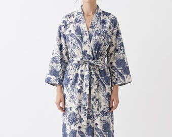Puede incluir: Una bata larga blanca con un estampado floral y de pájaros en azul. La bata tiene un diseño envolvente con un cinturón a juego. Las mangas son de tres cuartos y la bata está hecha de una tela ligera.