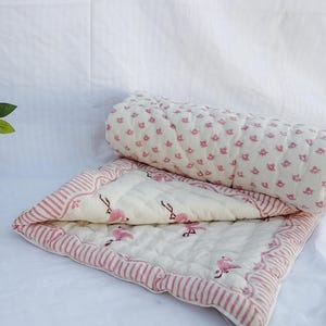 May include: A baby quilt featuring a flamingo design in pink and white. The quilt has a striped edge and is partly rolled. A terracotta pot with flowers is in the background.