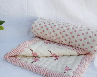 Puede incluir: Una colcha acolchada para bebé con un estampado de flamencos en rosa y blanco. La colcha tiene un borde a rayas y está parcialmente enrollada. Una maceta de terracota con flores está en el fondo.