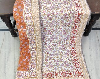 Puede incluir: Un edredón con estampado floral con fondo blanco y diseños florales rojos y dorados. El edredón tiene un borde con un patrón floral repetido y una sección naranja sólida en el lado izquierdo. El edredón está doblado sobre una mesa de madera.