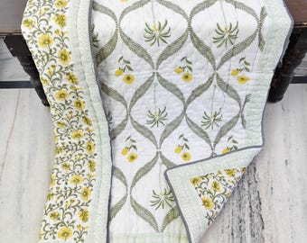 Puede incluir: Un edredón blanco con un patrón repetido de hojas verdes y flores amarillas. El borde presenta un diseño floral en amarillo y verde. El edredón está doblado sobre una superficie de madera oscura.