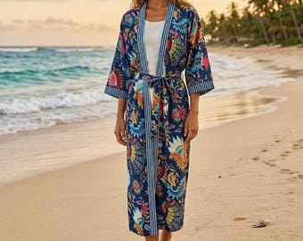 Puede incluir: Una mujer con un kimono largo azul marino con un colorido estampado floral y de pájaros, de pie en una playa de arena. El kimono tiene un ribete a rayas y un cinturón a juego. El fondo presenta el océano, palmeras y una puesta de sol.