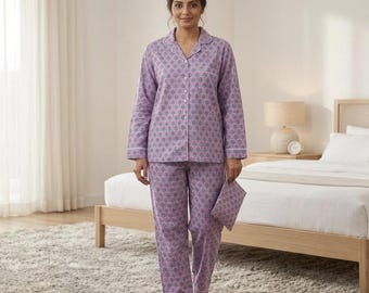 Puede incluir: Un conjunto de pijama lila con un estampado floral repetido. La camisa abotonada de manga larga y los pantalones a juego tienen ribetes blancos. La persona sostiene una máscara para dormir a juego. El pijama está hecho de un material suave y cómodo.