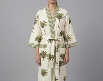 Puede incluir: Una bata color crema con un estampado de palmeras. La bata tiene ribetes verdes y un cinturón a juego. La bata llega a la rodilla y tiene bolsillos. Las palmeras son verdes y marrones.