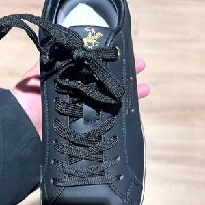 Può includere: Sneaker in pelle nera con lacci neri. La scarpa presenta un'emblema dorato e la scritta "Beverly Hills Polo Club" sul lato. La suola è bianca e la scarpa è nuova.