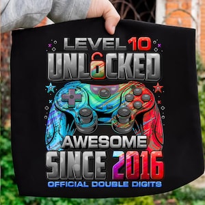 May include: Black graphic tee with a colorful video game controller design. Text includes "LEVEL 10 UNLOCKED AWESOME SINCE 2016 OFFICIAL DOUBLE DIGITS". The controller is blue, green, and red.