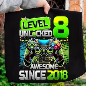 Könnte beinhalten: Schwarzes T-Shirt mit einem farbenfrohen Videospiel-Controller-Design. Das Shirt zeigt den Text "LEVEL UNLOCKED 8 AWESOME SINCE 2018" in Grün, Gelb und Weiß. Der Controller hat blaue, grüne, rote und gelbe Knöpfe.