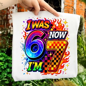 May include: White tote bag with a vibrant graphic. The text reads "I WAS 6 NOW I'M 7". The design includes a large number 6 with a lightning effect, a checkered number 7, and flames in a bright color palette.
