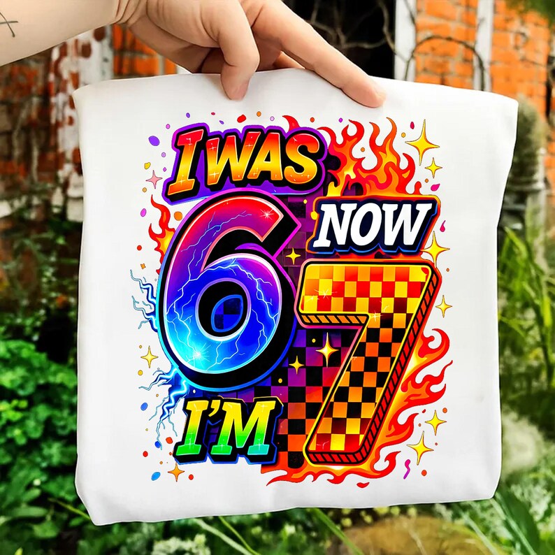 May include: White tote bag with a vibrant graphic. The text reads "I WAS 6 NOW I'M 7". The design includes a large number 6 with a lightning effect, a checkered number 7, and flames in a bright color palette.