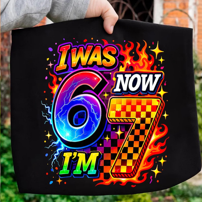 May include: Black graphic tee with the text "I WAS 6 NOW I'M 7". The number 6 is blue and purple with lightning bolts, and the number 7 is a checkered pattern with flames. The text is in various colours.