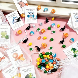 Pode incluir: Uma coleção de pequenos amuletos coloridos em forma de animais e cartões com mensagens positivas. Os amuletos incluem tartarugas, patos, leões, tubarões e corujas. Os cartões têm frases como "You're Awesome" e "Thanks for Owl You Do."