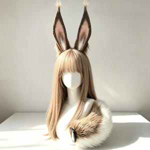 Könnte beinhalten: Eine lange, glatte, hellbraune Perücke mit Pony, die mit hohen, spitzen, pelzigen Tierohren in Braun-, Beige- und Weißtönen versehen ist. Ein passender, dicker, flauschiger, geschwungener Tierschwanz mit braunem Fell und weißer Spitze liegt an der Basis.