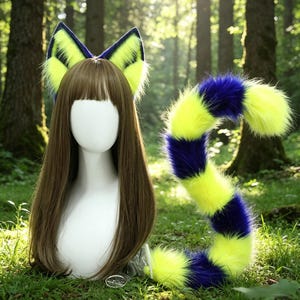 Puede incluir: Un conjunto de accesorios de cosplay con orejas de gato y una cola. Las orejas son de color amarillo neón y azul marino, y la cola está a rayas en los mismos colores. Los accesorios se muestran en una cabeza de maniquí con pelo castaño.