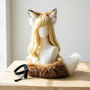 May include: A fox ear headband and tail set. The ears are brown, white, and black, and the tail is brown and white. The set is displayed on a mannequin head with blonde hair. The tail has a black ribbon.
