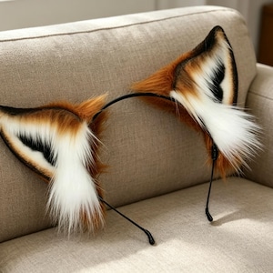 May include: A pair of fox ears on a black headband. The ears are a mix of brown, white, and black faux fur, with a fluffy texture. The headband is resting on a beige sofa.