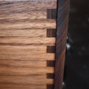 Puede incluir: Primer plano de una caja de madera con uniones de cola de milano. La caja est&aacute; hecha de madera marr&oacute;n claro con un borde marr&oacute;n m&aacute;s oscuro. Las uniones de cola de milano son visibles, mostrando la artesan&iacute;a de la carpinter&iacute;a.