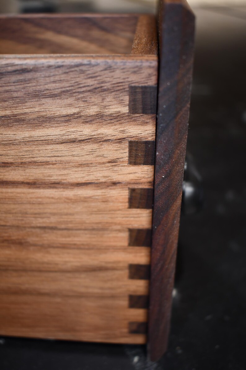 Puede incluir: Primer plano de una caja de madera con uniones de cola de milano. La caja est&aacute; hecha de madera marr&oacute;n claro con un borde marr&oacute;n m&aacute;s oscuro. Las uniones de cola de milano son visibles, mostrando la artesan&iacute;a de la carpinter&iacute;a.