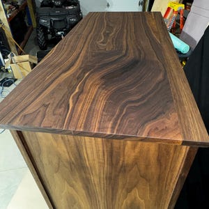 Puede incluir: Mesa de madera artesanal con acabado en nogal oscuro. La superficie superior muestra un patr&oacute;n de vetas de madera prominente, mientras que los lados tienen un tono de madera m&aacute;s claro y natural. El dise&ntilde;o es simple y moderno, mostrando la belleza natural de la madera.