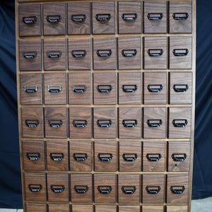 Puede incluir: Un mueble de boticario alto de madera con numerosos cajones peque&ntilde;os. El mueble es de madera oscura y tiene un solo caj&oacute;n grande en la parte superior. Cada caj&oacute;n peque&ntilde;o tiene un tirador de metal negro. El mueble est&aacute; dise&ntilde;ado para almacenamiento.