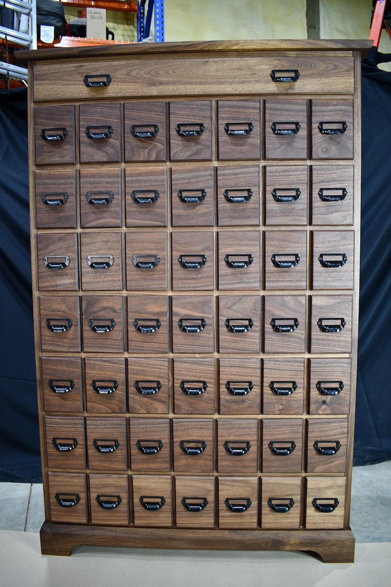 Puede incluir: Un mueble de boticario alto de madera con numerosos cajones peque&ntilde;os. El mueble es de madera oscura y tiene un solo caj&oacute;n grande en la parte superior. Cada caj&oacute;n peque&ntilde;o tiene un tirador de metal negro. El mueble est&aacute; dise&ntilde;ado para almacenamiento.