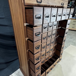 Puede incluir: Un mueble de boticario alto de madera con numerosos cajones peque&ntilde;os. El mueble es de madera marr&oacute;n oscuro y presenta tiradores de metal negro en cada caj&oacute;n. Los cajones est&aacute;n dispuestos en un patr&oacute;n de cuadr&iacute;cula, con algunos cajones abiertos. El mueble tiene una est&eacute;tica cl&aacute;sica y vintage.