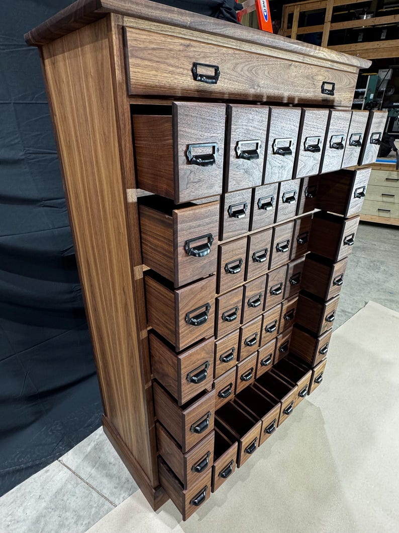 Puede incluir: Un mueble de boticario alto de madera con numerosos cajones peque&ntilde;os. El mueble es de madera marr&oacute;n oscuro y presenta tiradores de metal negro en cada caj&oacute;n. Los cajones est&aacute;n dispuestos en un patr&oacute;n de cuadr&iacute;cula, con algunos cajones abiertos. El mueble tiene una est&eacute;tica cl&aacute;sica y vintage.