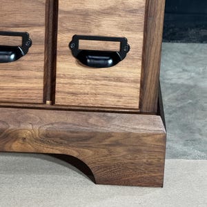 Puede incluir: Primer plano de un mueble de madera con varios cajones. El mueble es de madera marr&oacute;n oscuro con vetas visibles. Cada caj&oacute;n tiene un tirador de metal negro. El mueble se apoya sobre una base curva.
