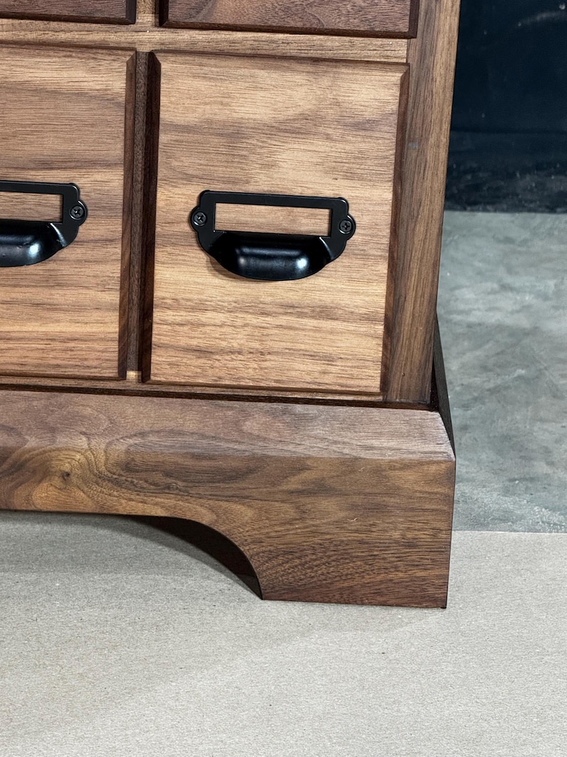 Puede incluir: Primer plano de un mueble de madera con varios cajones. El mueble es de madera marr&oacute;n oscuro con vetas visibles. Cada caj&oacute;n tiene un tirador de metal negro. El mueble se apoya sobre una base curva.