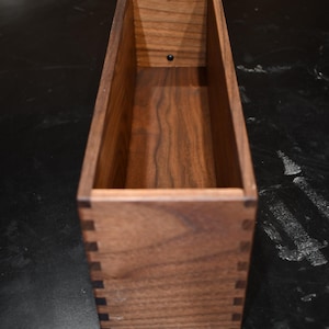 Puede incluir: Una caja rectangular de madera con juntas de cola de milano y un acabado marr&oacute;n oscuro. La caja tiene una abertura rectangular y est&aacute; construida con madera maciza. El interior es visible, mostrando la veta de la madera. La caja est&aacute; vac&iacute;a.