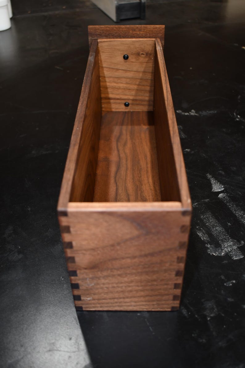 Puede incluir: Una caja rectangular de madera con juntas de cola de milano y un acabado marr&oacute;n oscuro. La caja tiene una abertura rectangular y est&aacute; construida con madera maciza. El interior es visible, mostrando la veta de la madera. La caja est&aacute; vac&iacute;a.