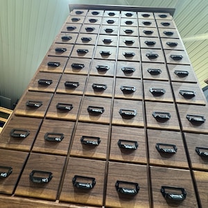 May include: A tall, wooden cabinet with numerous drawers. The cabinet is constructed from dark brown wood and features rows of small, square drawers with black metal handles. The drawers are arranged in a grid pattern, creating a visually appealing storage unit.