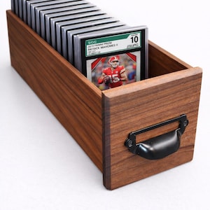 Puede incluir: Una caja de almacenamiento de tarjetas de madera con un asa negra, llena de tarjetas deportivas clasificadas. La tarjeta superior presenta a Patrick Mahomes II. Las tarjetas est&aacute;n en estuches protectores. La caja es de madera oscura.
