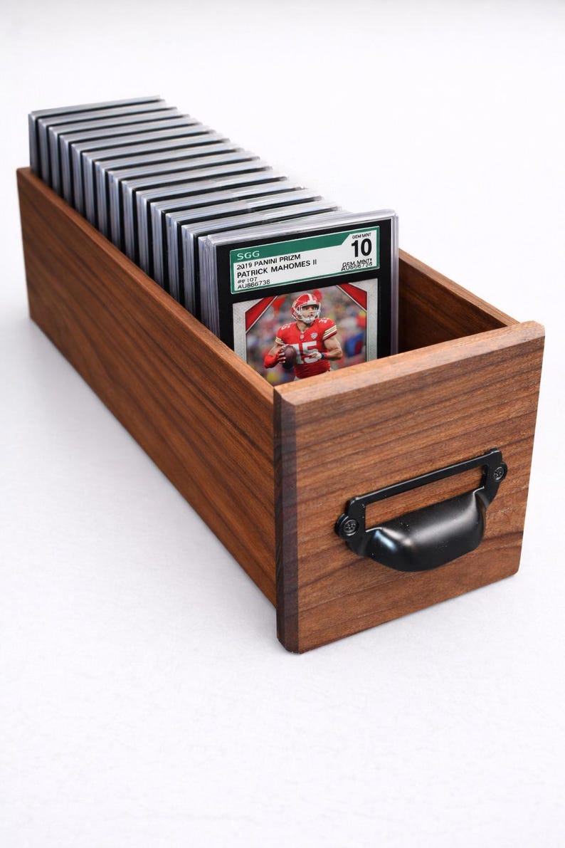 Puede incluir: Una caja de almacenamiento de tarjetas de madera con un asa negra, llena de tarjetas deportivas clasificadas. La tarjeta superior presenta a Patrick Mahomes II. Las tarjetas est&aacute;n en estuches protectores. La caja es de madera oscura.