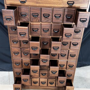 Puede incluir: Un mueble de boticario de madera oscura con numerosos cajones peque&ntilde;os, algunos abiertos. El mueble tiene tiradores de metal negro. Los cajones est&aacute;n dispuestos en un patr&oacute;n de cuadr&iacute;cula, creando una soluci&oacute;n de almacenamiento visualmente atractiva.