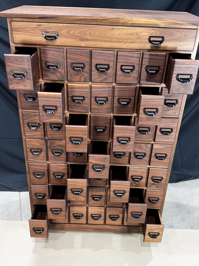 Puede incluir: Un mueble de boticario de madera oscura con numerosos cajones peque&ntilde;os, algunos abiertos. El mueble tiene tiradores de metal negro. Los cajones est&aacute;n dispuestos en un patr&oacute;n de cuadr&iacute;cula, creando una soluci&oacute;n de almacenamiento visualmente atractiva.