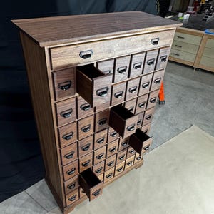 Puede incluir: Un mueble de boticario alto de madera con numerosos cajones peque&ntilde;os. El mueble es de madera oscura y tiene tiradores met&aacute;licos en cada caj&oacute;n. Varios cajones est&aacute;n abiertos, revelando el interior.