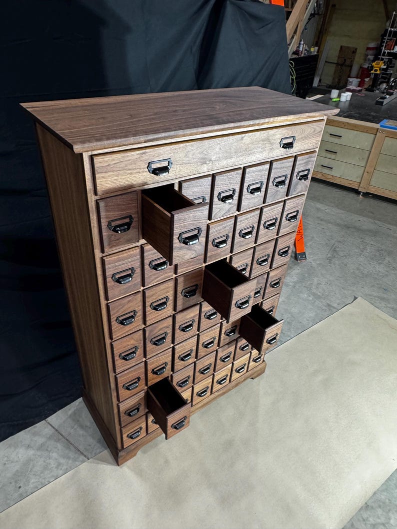 Puede incluir: Un mueble de boticario alto de madera con numerosos cajones peque&ntilde;os. El mueble es de madera oscura y tiene tiradores met&aacute;licos en cada caj&oacute;n. Varios cajones est&aacute;n abiertos, revelando el interior.