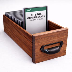 Puede incluir: Una caja de almacenamiento de tarjetas de madera con un asa negra. La caja contiene tarjetas clasificadas, con una tarjeta visible, que muestra el texto "FITS SGC GRADED CARDS AND SIMILAR SLABS". La caja es de madera con veta visible.