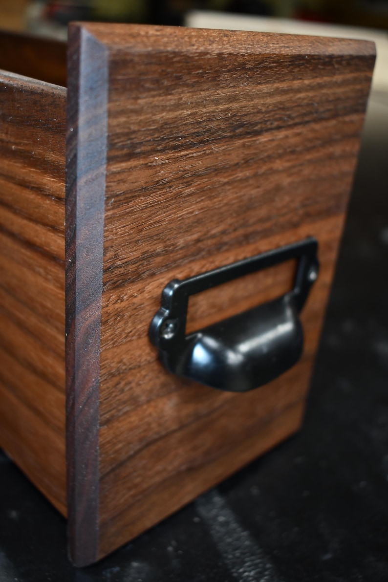 Puede incluir: Primer plano de una caja de madera con acabado oscuro y un asa de metal negro. La caja tiene forma rectangular con veta de madera visible. El asa est&aacute; fijada a la parte delantera de la caja.