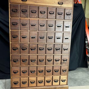 Puede incluir: Un mueble de boticario de madera con numerosos cajones peque&ntilde;os, cada uno con un tirador de metal. El mueble es de madera oscura y tiene forma rectangular. Los cajones est&aacute;n dispuestos en forma de rejilla y el mueble tiene un caj&oacute;n superior.